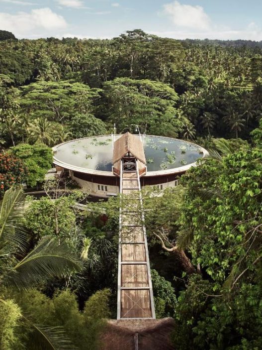 An aerial view of a large circular rooftop lotus pond and a wooden suspension bridge at Four Seasons Resort Bali at Sayan, surrounded by a dense tropical rainforest under a clear sky.