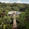 An aerial view of a large circular rooftop lotus pond and a wooden suspension bridge at Four Seasons Resort Bali at Sayan, surrounded by a dense tropical rainforest under a clear sky.