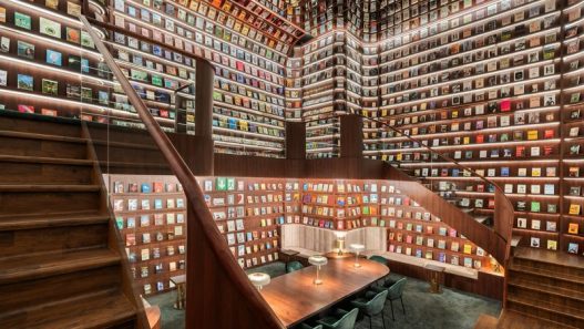 A grand, multi-level 'Secret Library' with floor-to-ceiling wooden bookshelves filled with colorful books. The room features a curved wooden staircase, warm LED shelf lighting, and a long wooden communal table with green velvet chairs.