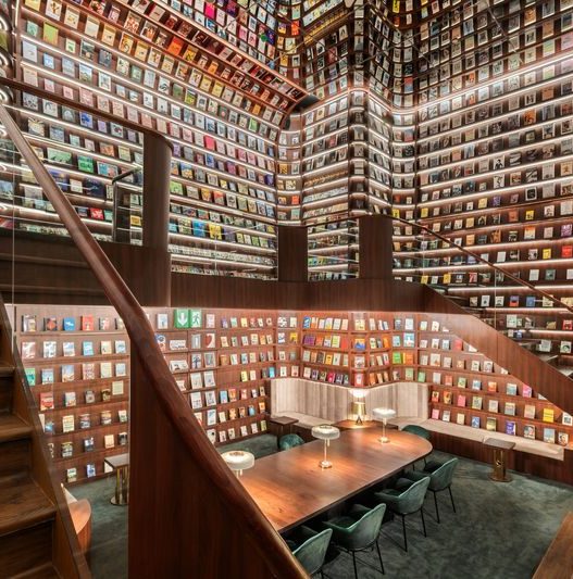 A grand, multi-level 'Secret Library' with floor-to-ceiling wooden bookshelves filled with colorful books. The room features a curved wooden staircase, warm LED shelf lighting, and a long wooden communal table with green velvet chairs.
