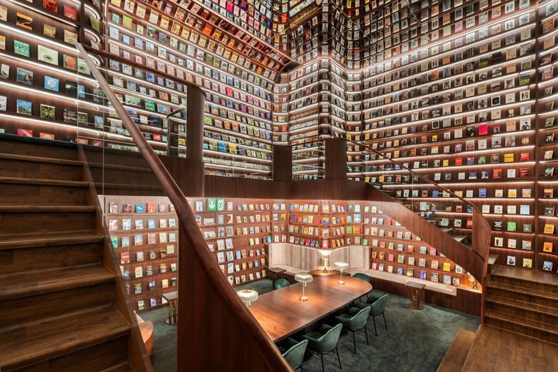 A grand, multi-level 'Secret Library' with floor-to-ceiling wooden bookshelves filled with colorful books. The room features a curved wooden staircase, warm LED shelf lighting, and a long wooden communal table with green velvet chairs.